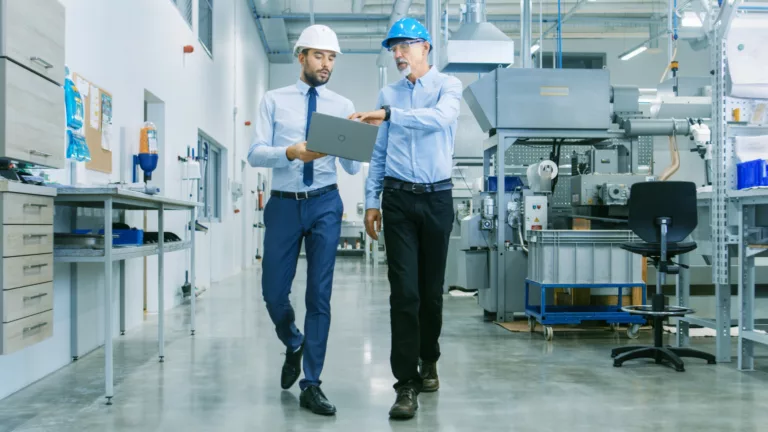 Two men walking in a clean manufacturing environment discussing something on a laptop computer.